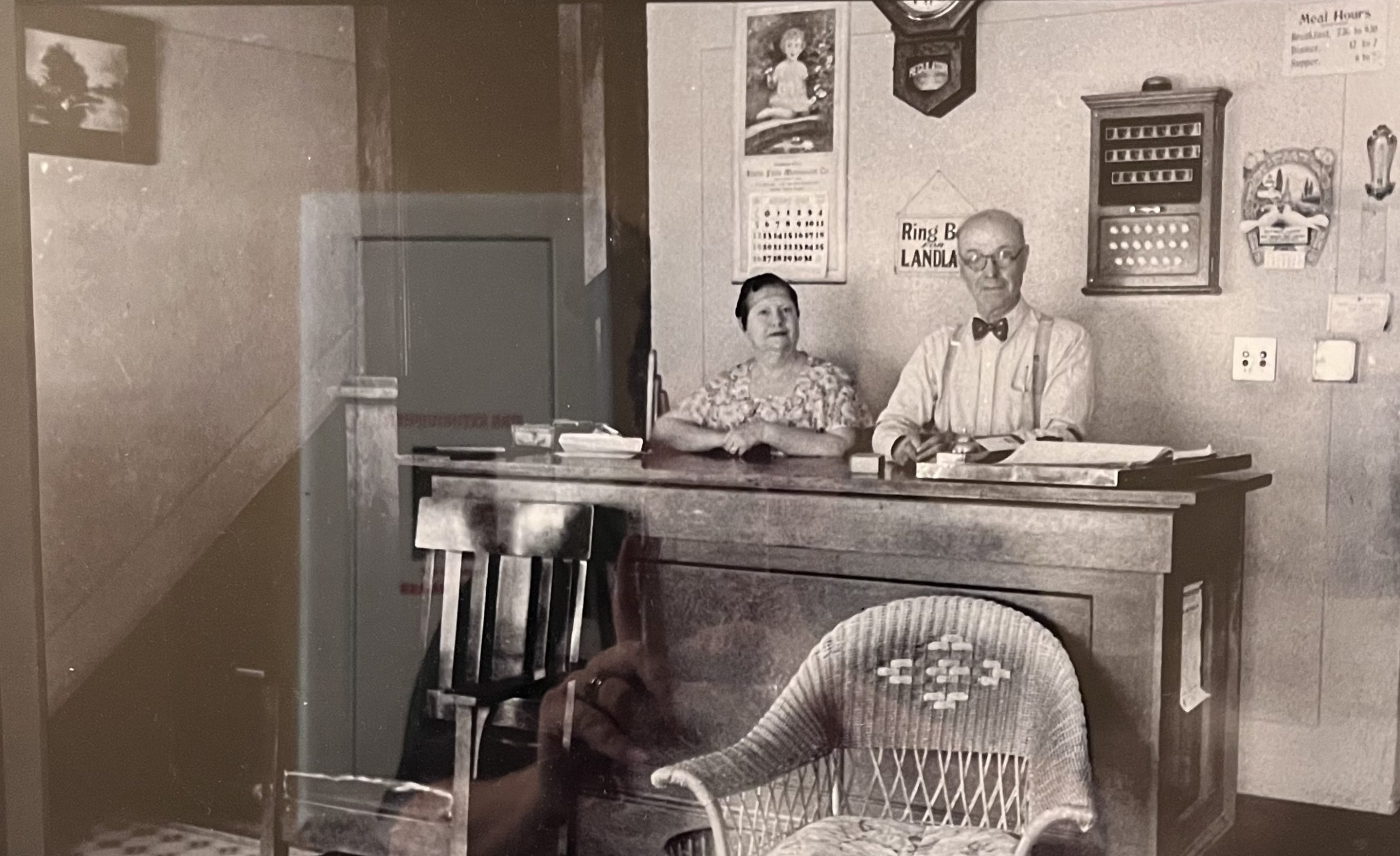 Black-and-white photo of a vintage hotel lobby with a man in a bow tie and a woman in a floral dress behind a wooden counter, exuding a nostalgic atmosphere.