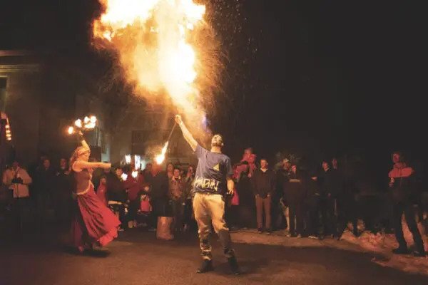 A fire performer breathes flames into the night sky, captivating a crowd gathered in a circle. Another performer dances nearby. The scene feels exciting and dramatic.