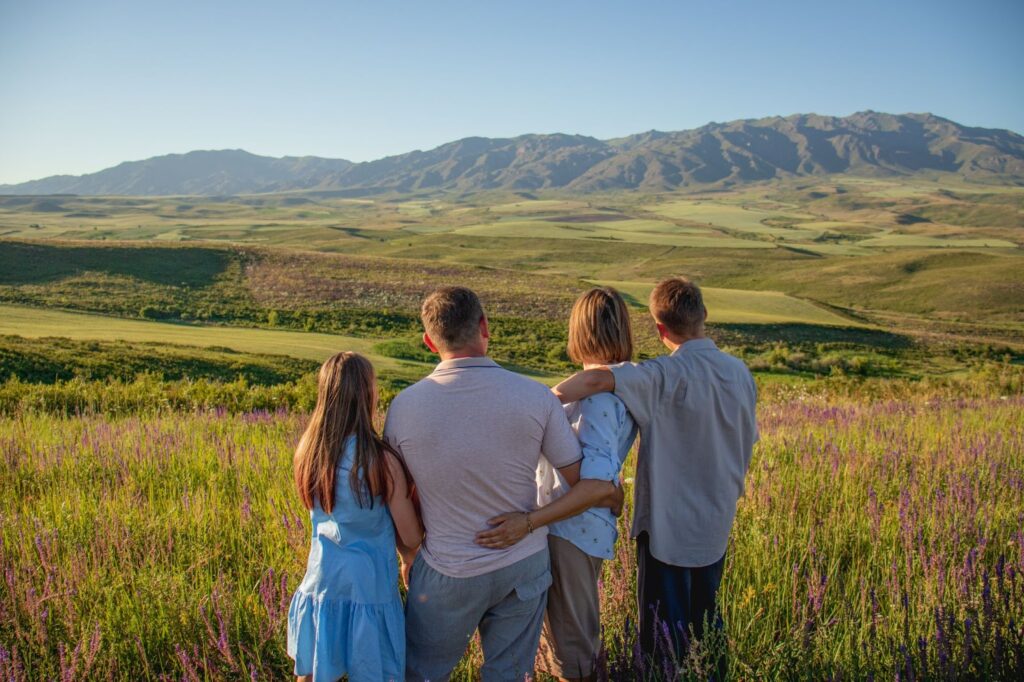A family of four standing together in a field of purple flowers, gazing at distant green hills under a clear blue sky.
