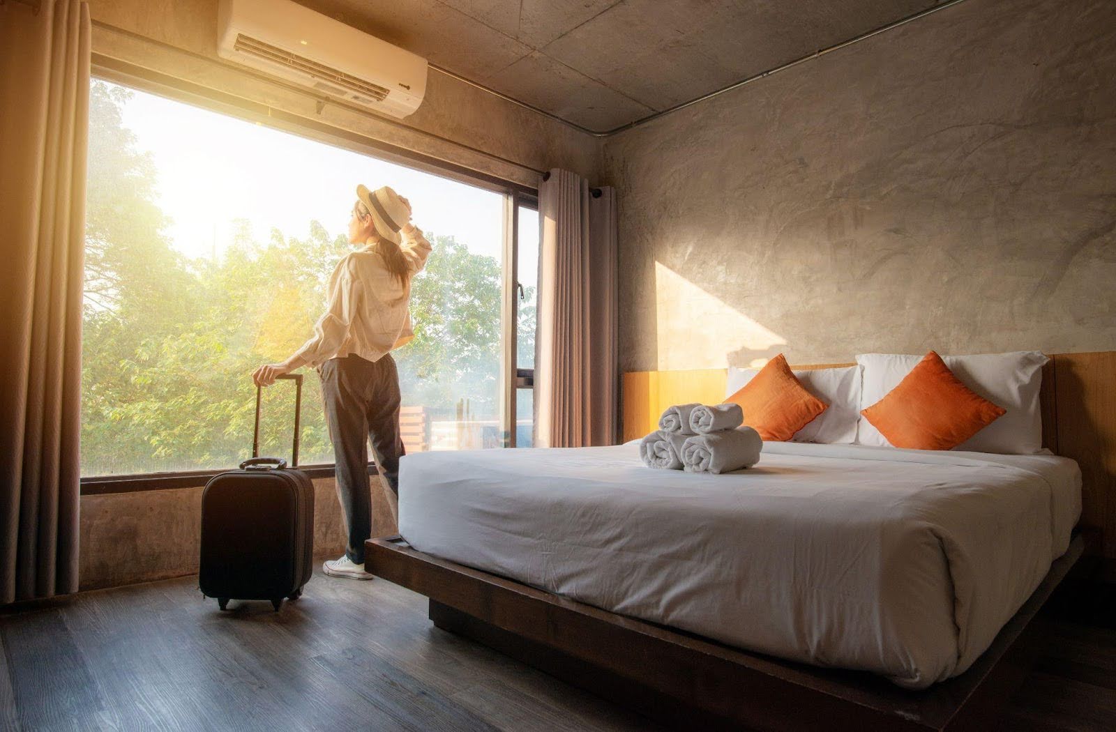 A woman stands in front of a bed next to a suitcase preparing for a trip