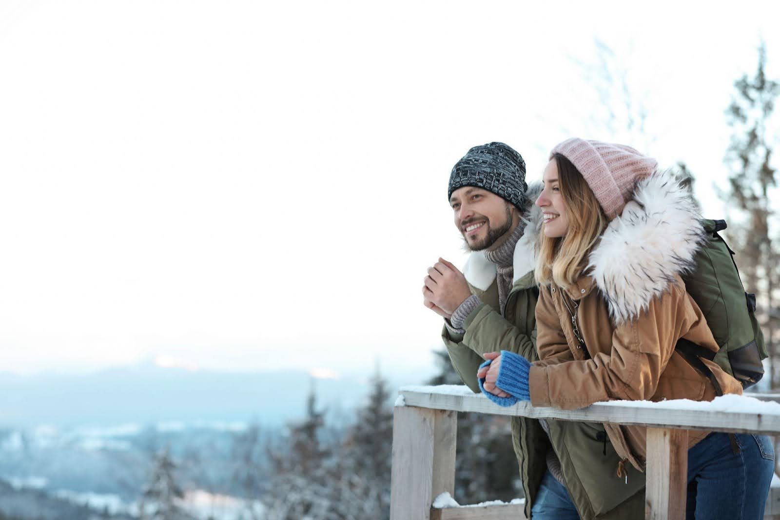 A man and woman stand together on a snowy hillside surrounded by a winter landscape