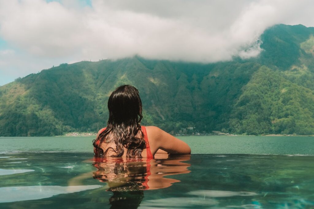 A woman with long dark hair, wearing a red swimsuit, relaxes in an infinity pool overlooking a serene turquoise lake with lush green mountains shrouded in clouds in the background.