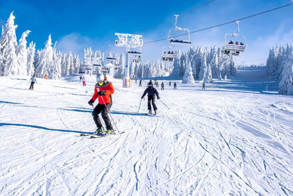 Skiers descending a groomed slope on a sunny day, with a chairlift overhead and snow-covered evergreen trees in the background.