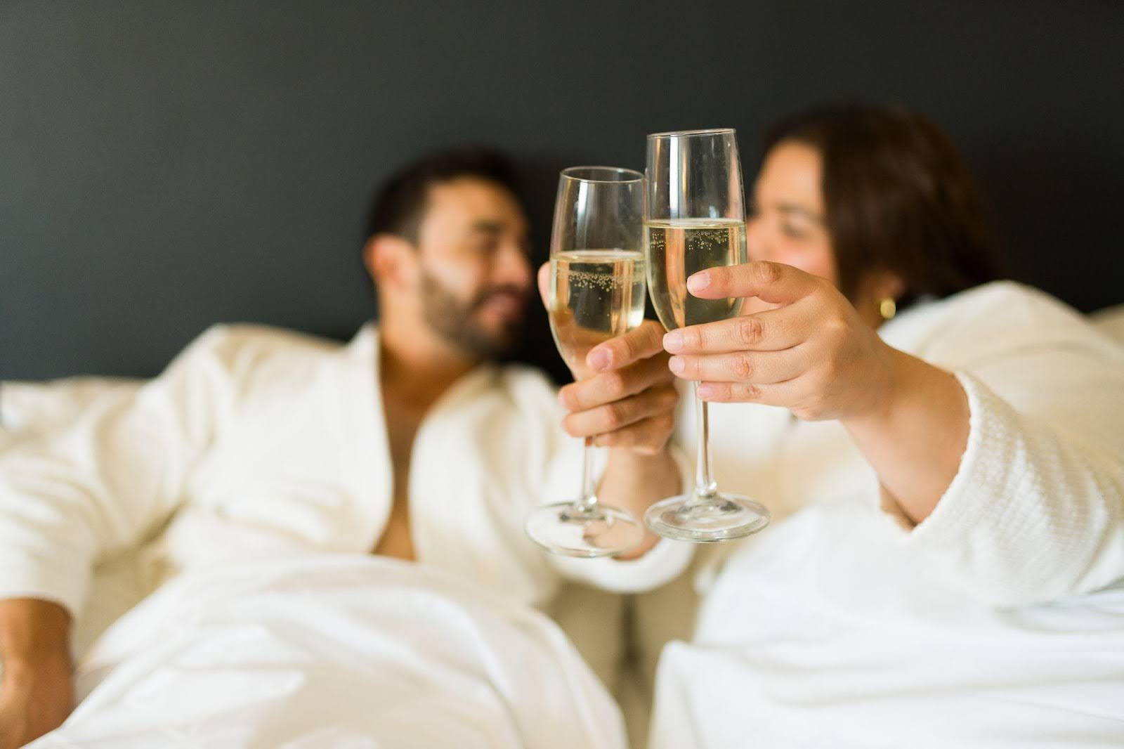 A couple in bed, raising champagne glasses in celebration with cozy bedding creating an intimate setting