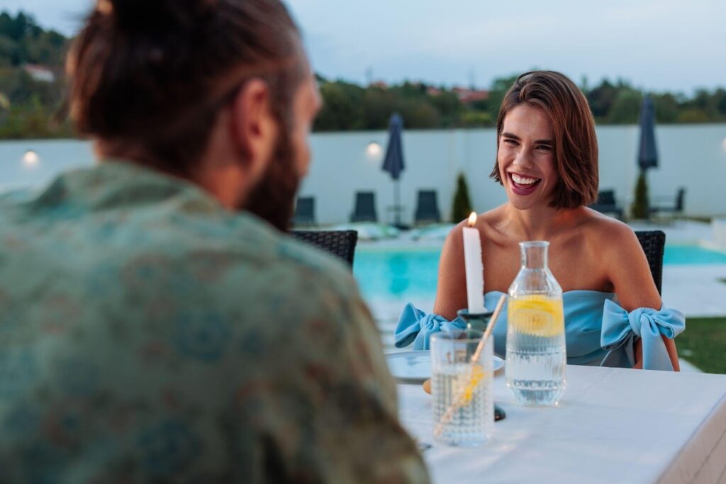 A man and woman are seated at a table next to a pool engaged in conversation under a sunny sky