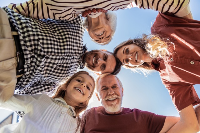 A low-angle view looking up at a multi-generational family of five smiling and huddling together in a circle against a bright blue sky.