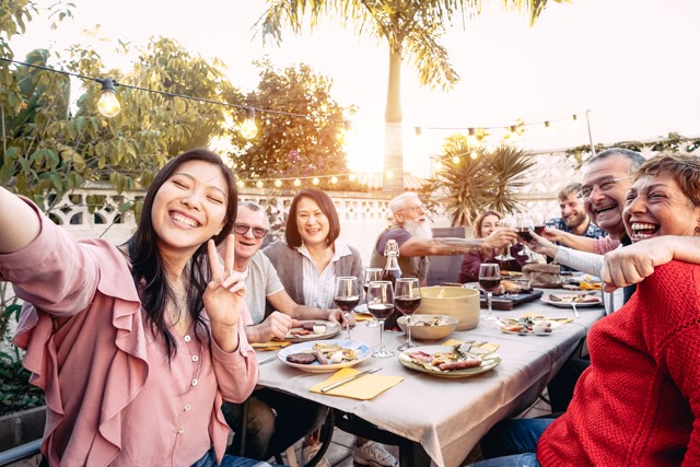 A smiling young woman takes a selfie with a diverse group of friends and family enjoying an outdoor dinner party at a long table under string lights.