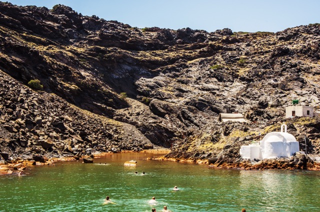 People swimming in the green waters of a volcanic bay in Santorini, Greece, with rugged dark cliffs and a small white Orthodox church on the shore.