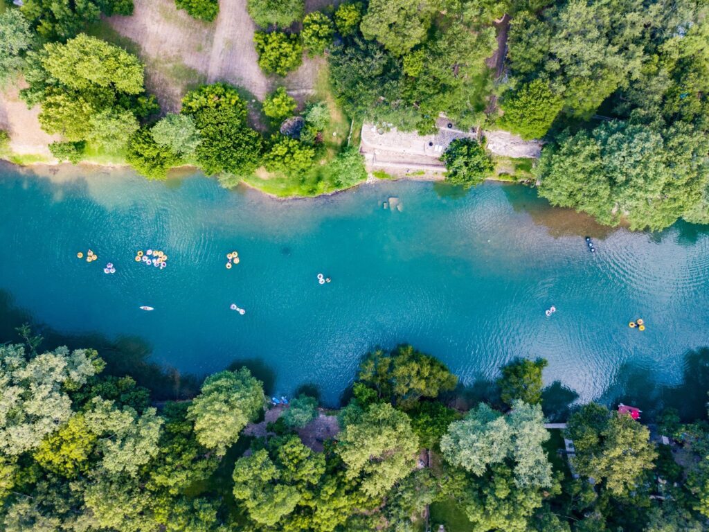 An aerial view shows numerous people floating in colorful inner tubes on a bright turquoise river lined with dense green trees.