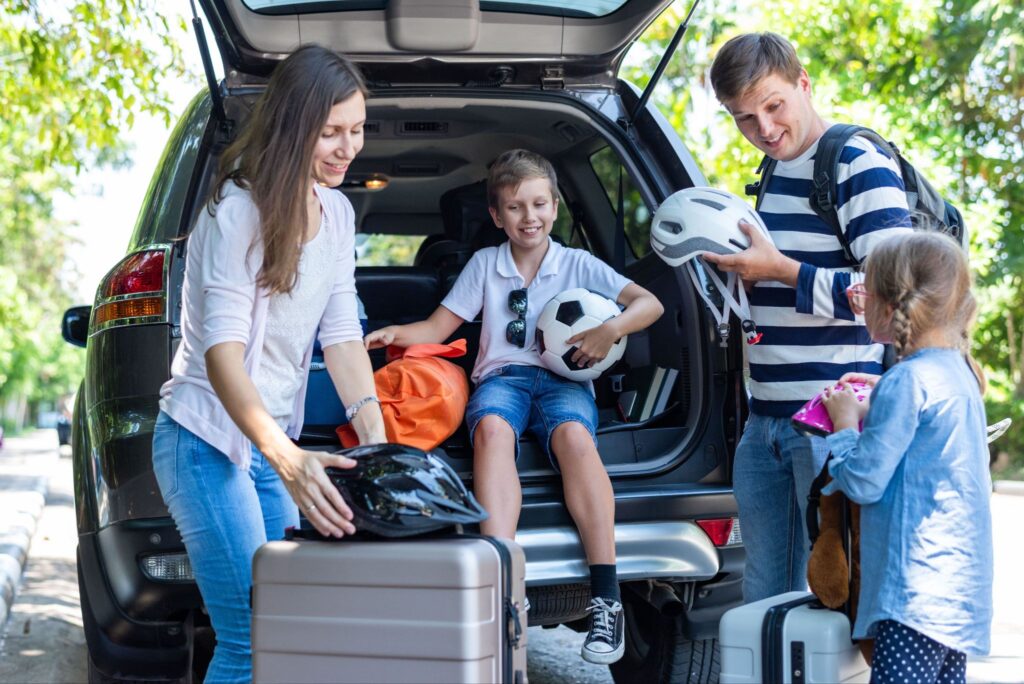 Family packing luggage, helmets, and a soccer ball into the trunk of a car for a road trip.