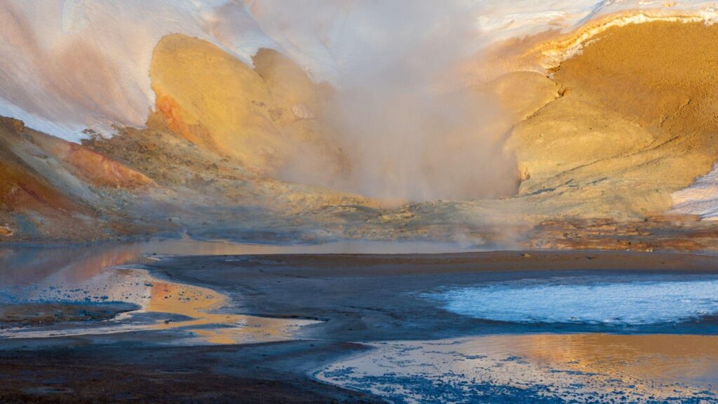 A large steaming geothermal hole surrounded by rocky terrain emitting vapor into the air