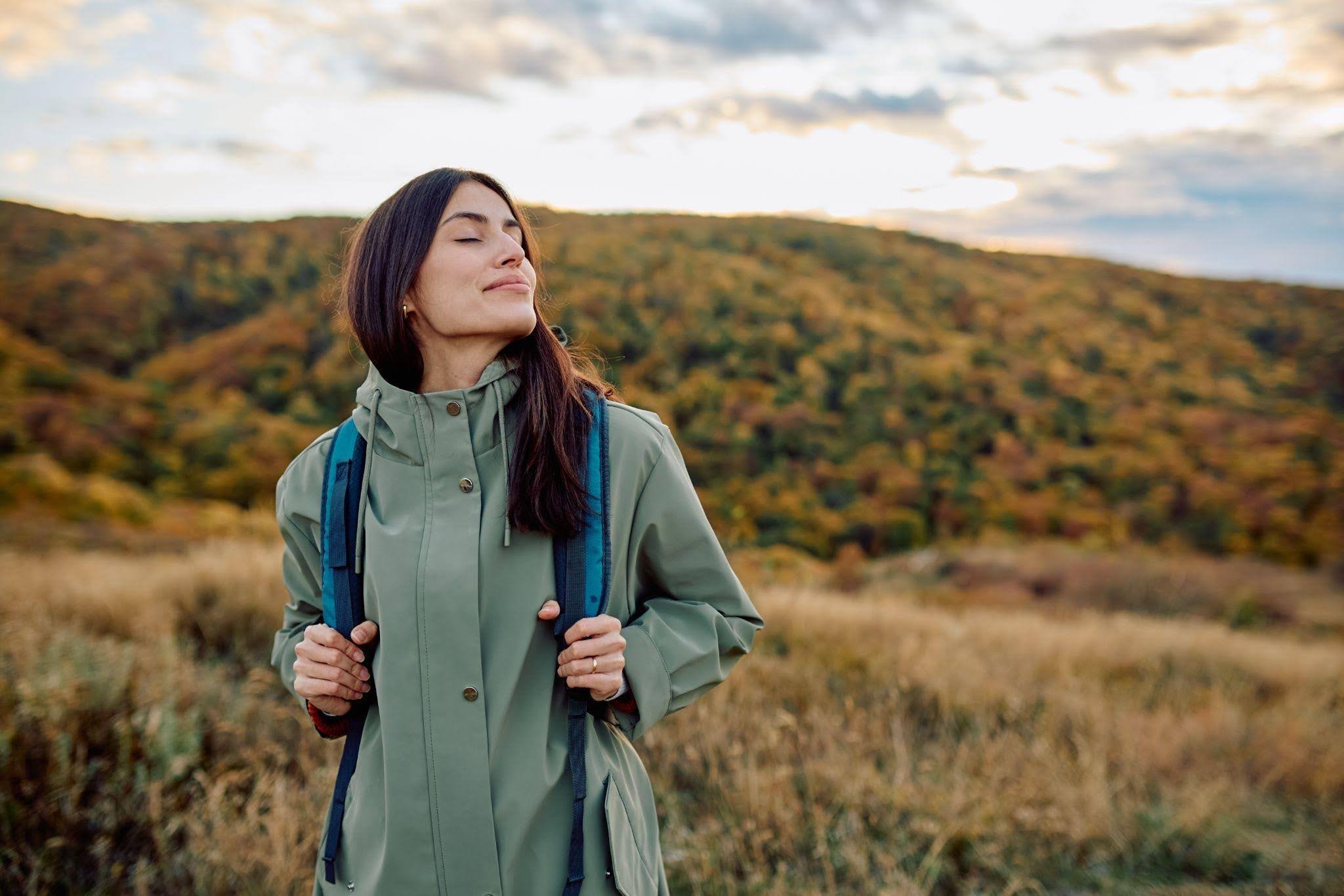 A woman wearing a green raincoat stands in a vast field surrounded by tall grass and a cloudy sky