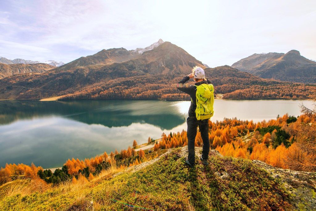A man stands on a mountain peak gazing at a serene lake surrounded by distant mountains